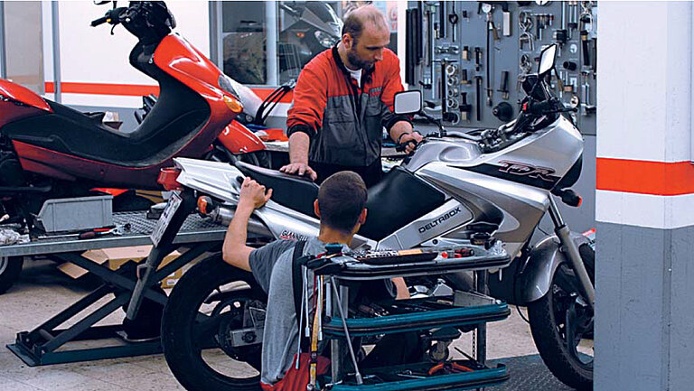 KFZ Werkstatt Bild einer Werkstatt, in der zwei Personen an einem Motorrad arbeiten.